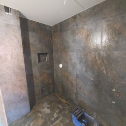 ProGlazurnik - Wnętrze łazienki w trakcie prac wykończeniowych, widoczne płytki imitujące kamień na ścianach, nieukończone podłączenia hydrauliczne i elektryczne, oraz narzędzia w kuwecie malarskiej na podłodze.