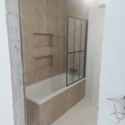 Wanna obudowana płytkami imitującymi drewno ułożonymi w jodełkę, z zamontowaną czarną kabiną prysznicową, w trakcie prac wykończeniowych.