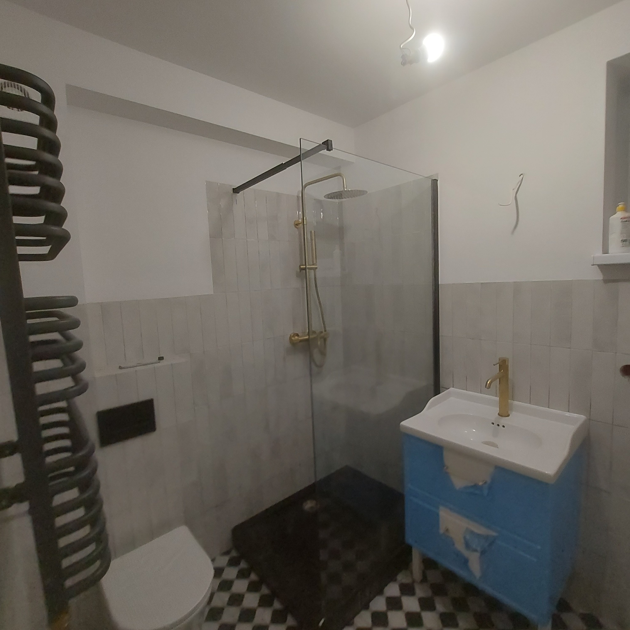 Zakończony remont łazienki: kabina prysznicowa typu walk-in z czarną ramą i złotą armaturą, niebieska szafka umywalkowa z uszkodzeniami folii, grzejnik drabinkowy i toaleta na tle biało-szarej...