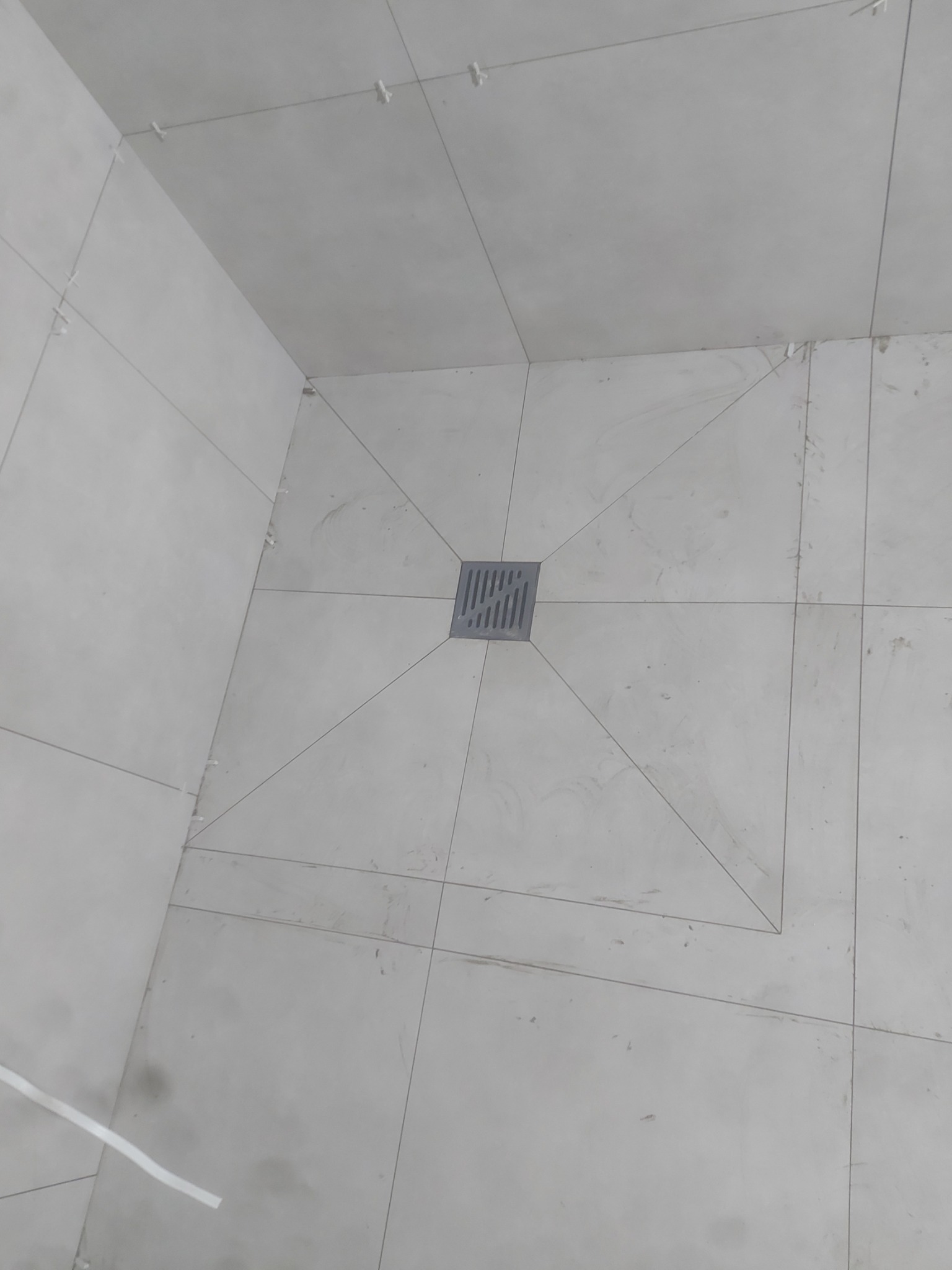 Świeżo ułożone, jasnoszare płytki podłogowe w kabinie prysznicowej z centralnie umieszczonym odpływem liniowym; widoczne krzyżyki dystansowe i ślady fugowania.
