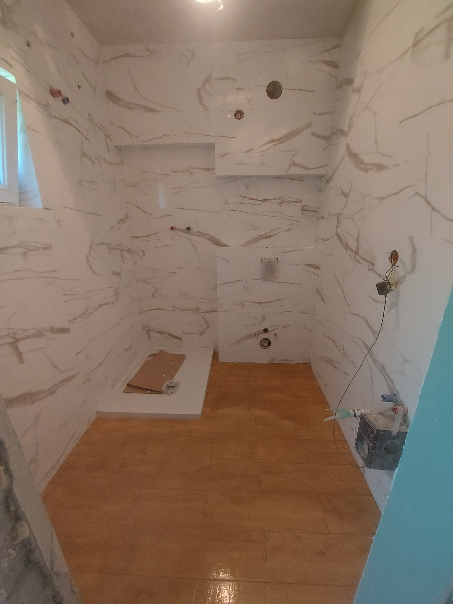 Wnętrze łazienki w trakcie prac wykończeniowych: ściany wyłożone białymi płytkami imitującymi marmur z widocznymi otworami na instalacje, brązowa podłoga z płytek, karton na podłodze w miejscu...