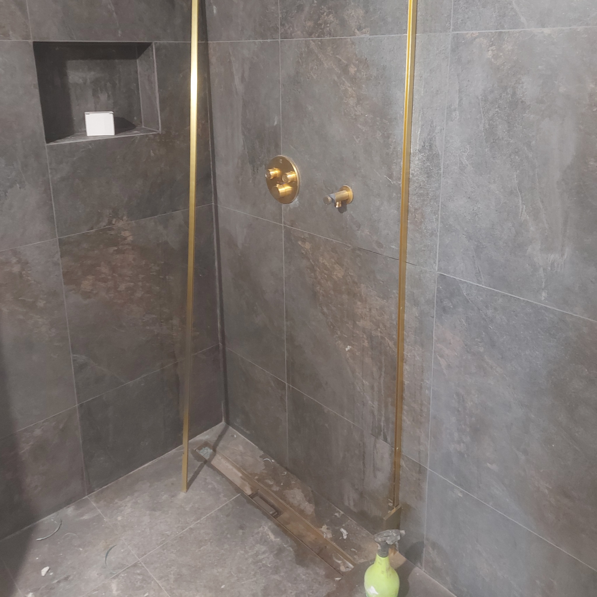 Szara łazienka z kabiną prysznicową ze złotymi elementami, wnęka z pudełkiem, widoczne ślady po montażu i butelka ze spryskiwaczem.