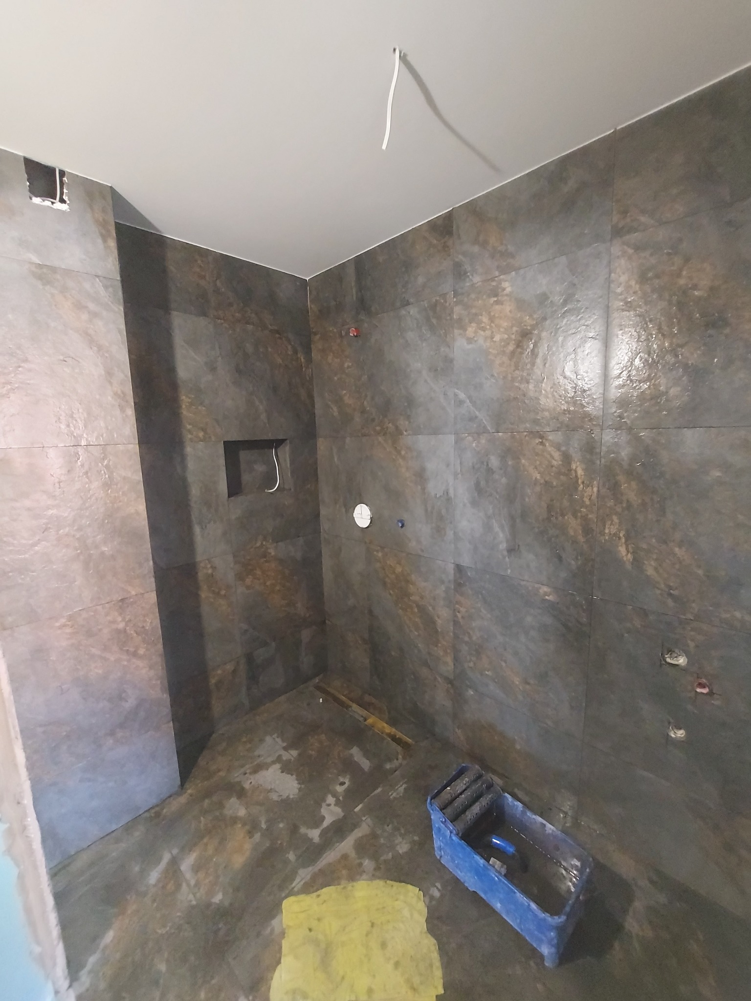 Wnętrze łazienki w trakcie prac wykończeniowych, widoczne płytki imitujące kamień na ścianach, nieukończone podłączenia hydrauliczne i elektryczne, oraz narzędzia w kuwecie malarskiej na podłodze.