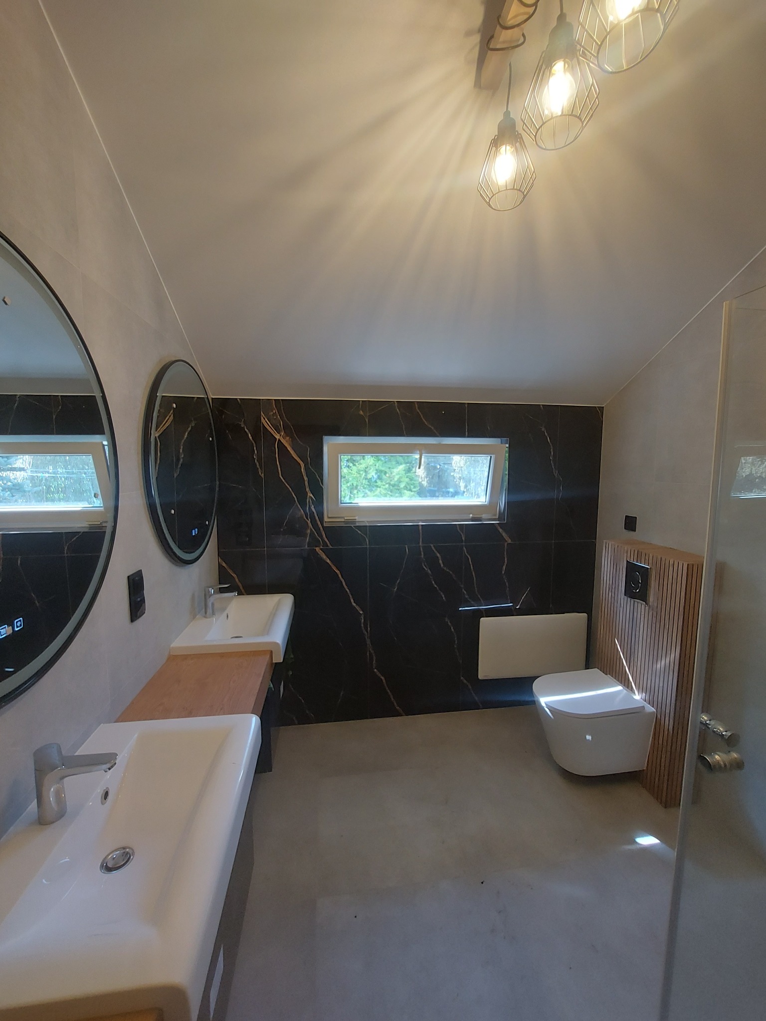 Nowoczesna łazienka z czarnymi płytkami imitującymi marmur, okrągłymi lustrami z podświetleniem LED, drewnianym blatem pod umywalkami i podwieszaną toaletą z drewnianą zabudową.