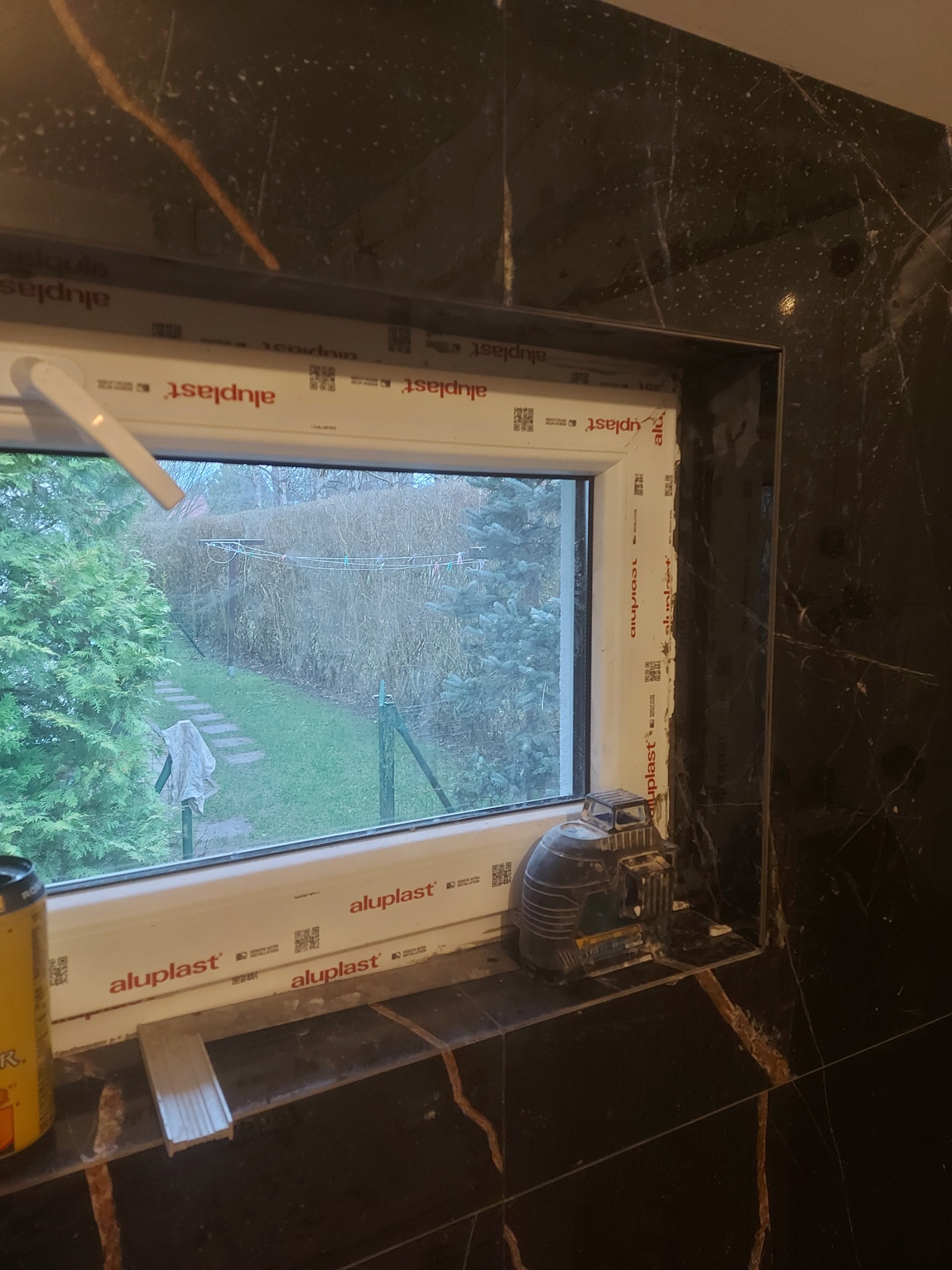 Okno PCV marki Aluplast osadzone w czarnych marmurowych płytkach podczas remontu, widoczny poziomica laserowa i puszka piwa.