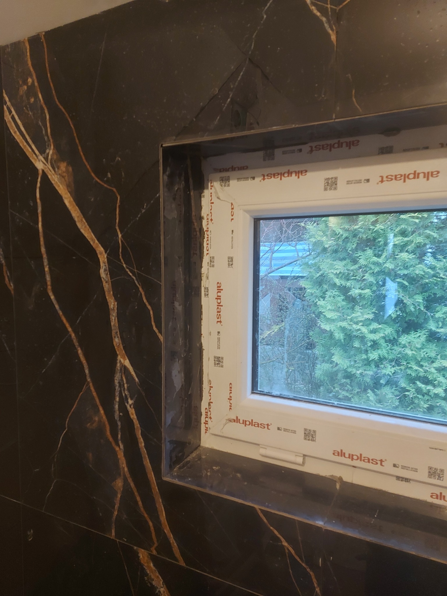 Czarna marmurowa okładzina ścienna z widocznym oknem PCV marki Aluplast i zielonymi drzewami za szybą.