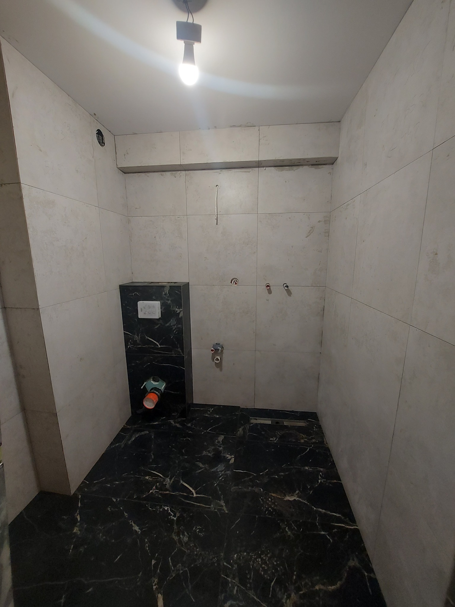 Niedokończona łazienka z szarymi płytkami ściennymi i czarną marmurową podłogą, widoczna podtynkowa toaleta i wystające rury instalacji wodnej.