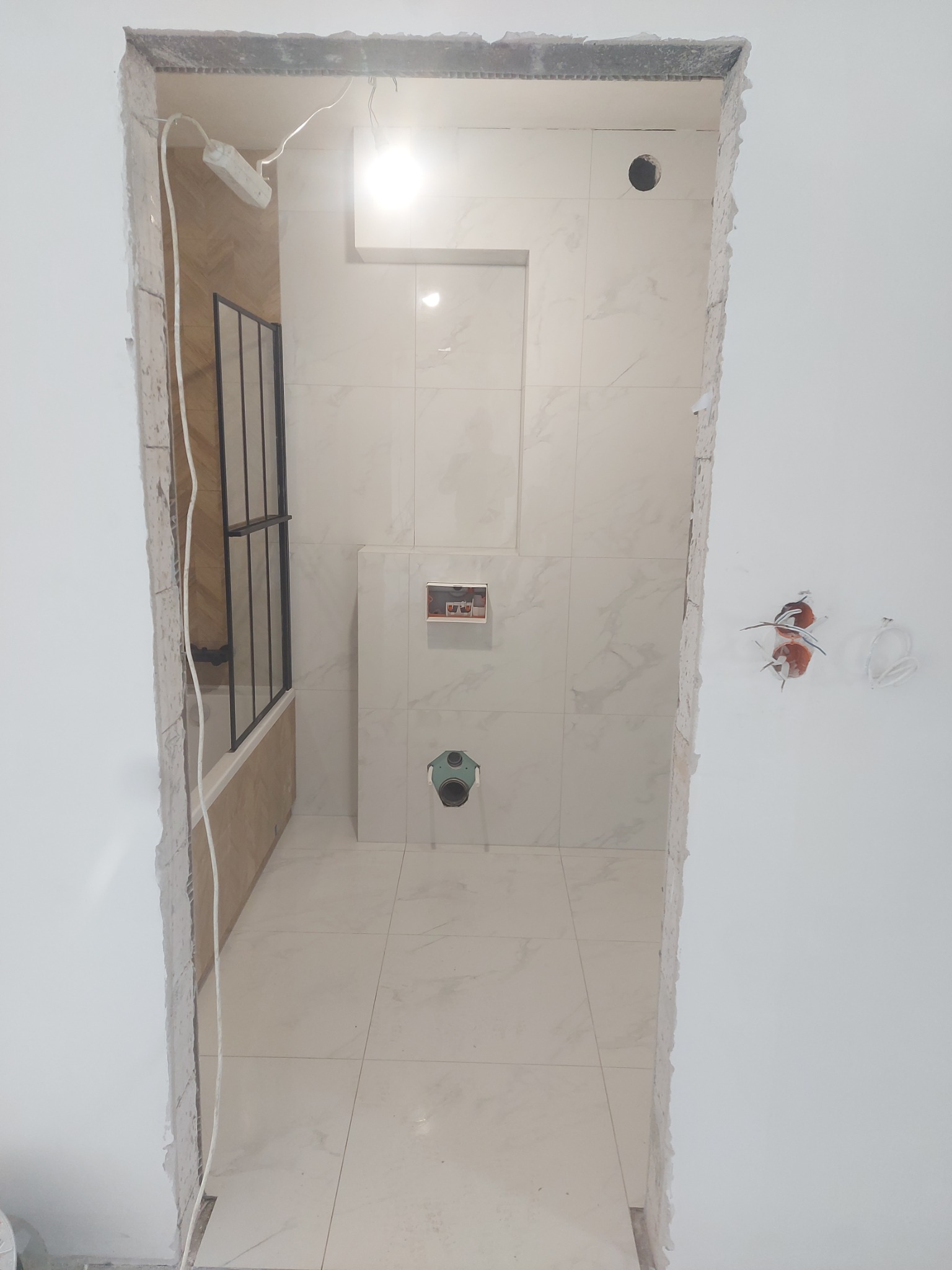 Łazienka w trakcie remontu: widoczne białe płytki imitujące marmur na ścianach i podłodze, fragment wanny z czarną kabiną, niedokończone ściany wokół wejścia, wystające kable elektryczne.