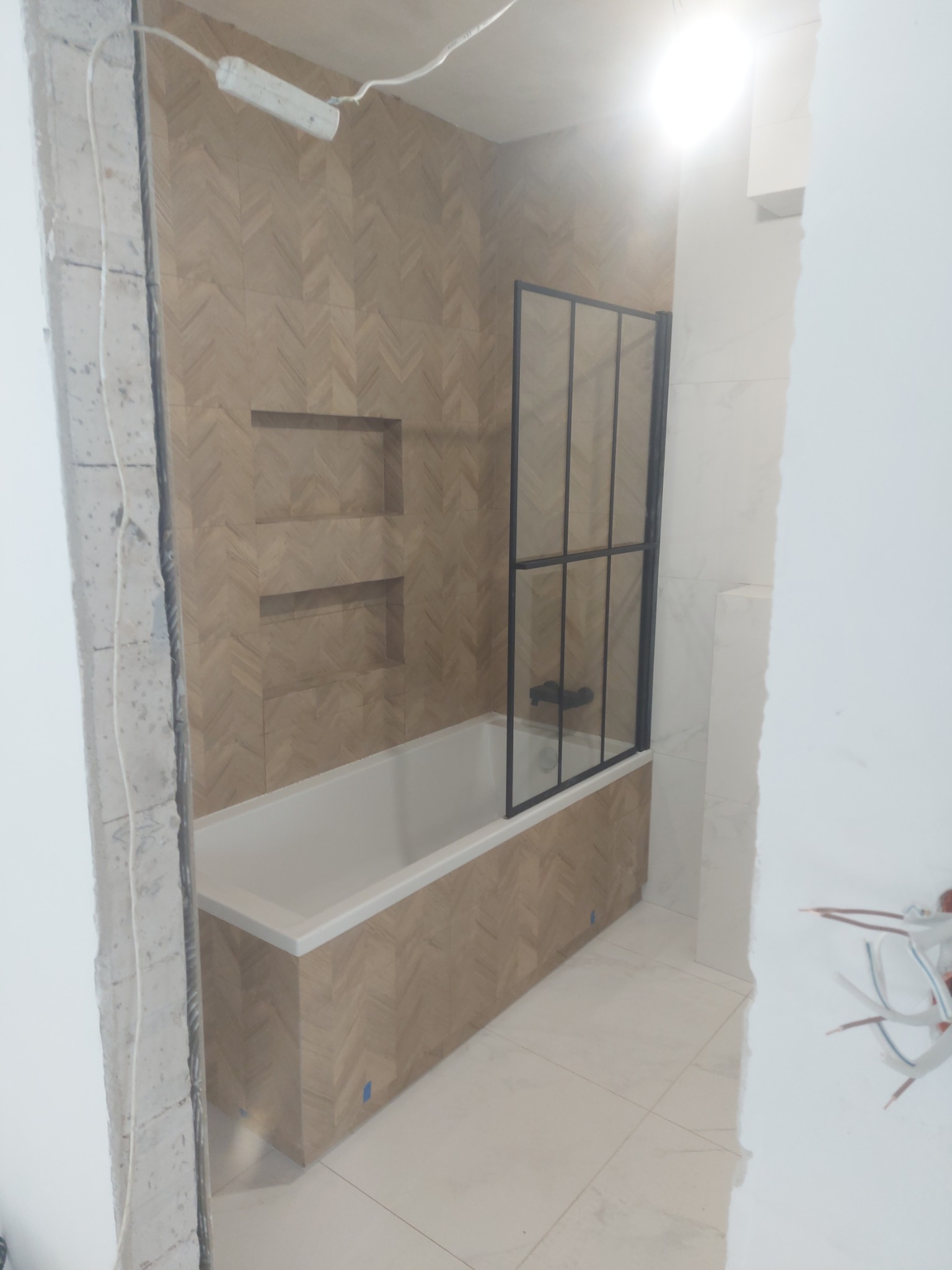 Wanna obudowana płytkami imitującymi drewno ułożonymi w jodełkę, z zamontowaną czarną kabiną prysznicową, w trakcie prac wykończeniowych.