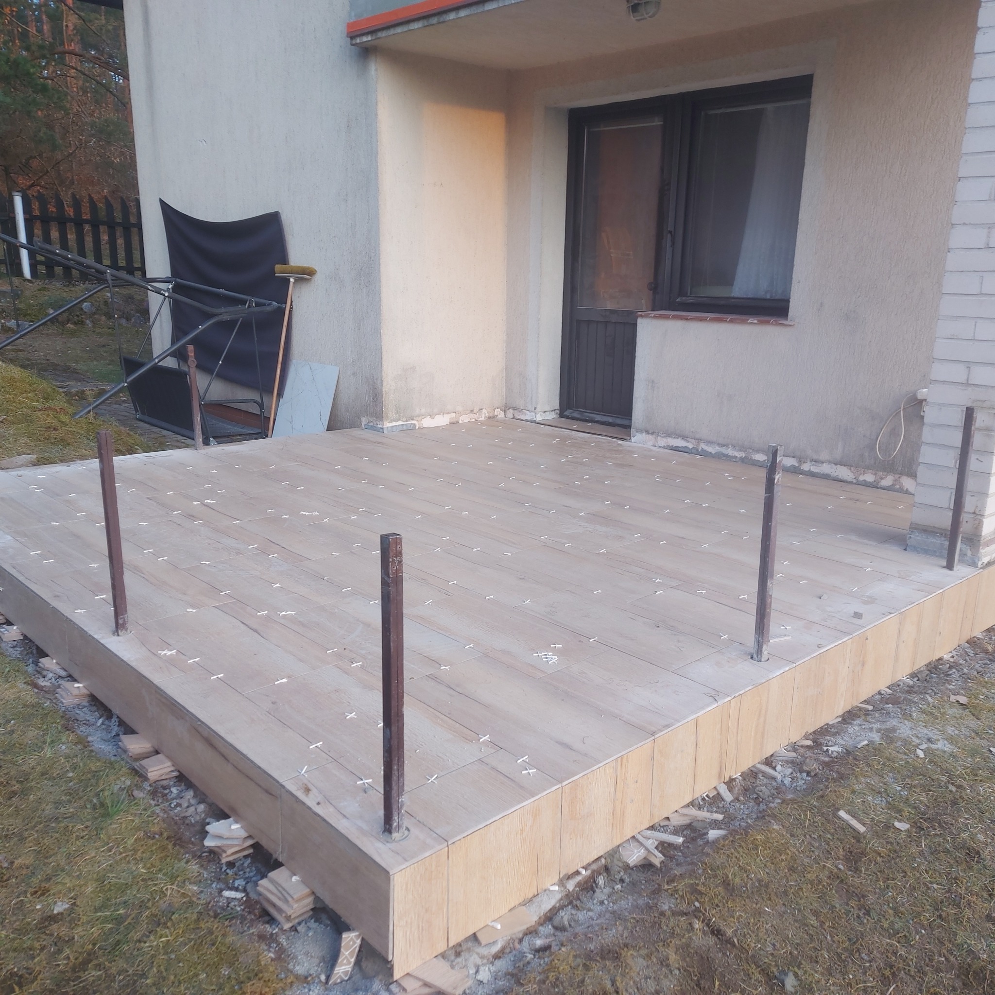 Układanie płytek imitujących drewno na tarasie, widoczne krzyżyki dystansowe, pionowe słupki i fragment elewacji budynku.