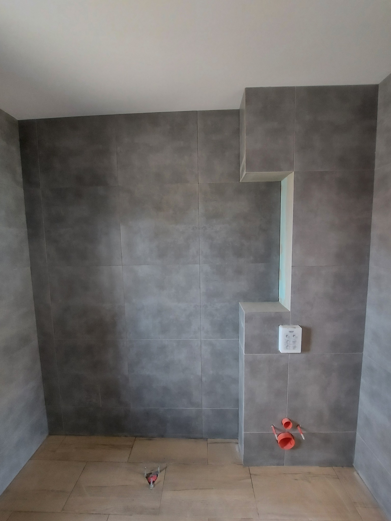 Ściany łazienki wyłożone szarymi płytkami imitującymi beton, z widoczną wnęką, instalacją wodną i częściowo wykończoną podłogą z jasnych płytek.