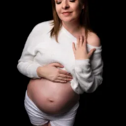 Kobieta w ciąży, w białym swetrze i koronkowych spodenkach, z delikatnym makijażem oczu, pozuje na czarnym tle, trzymając ręce na brzuchu.