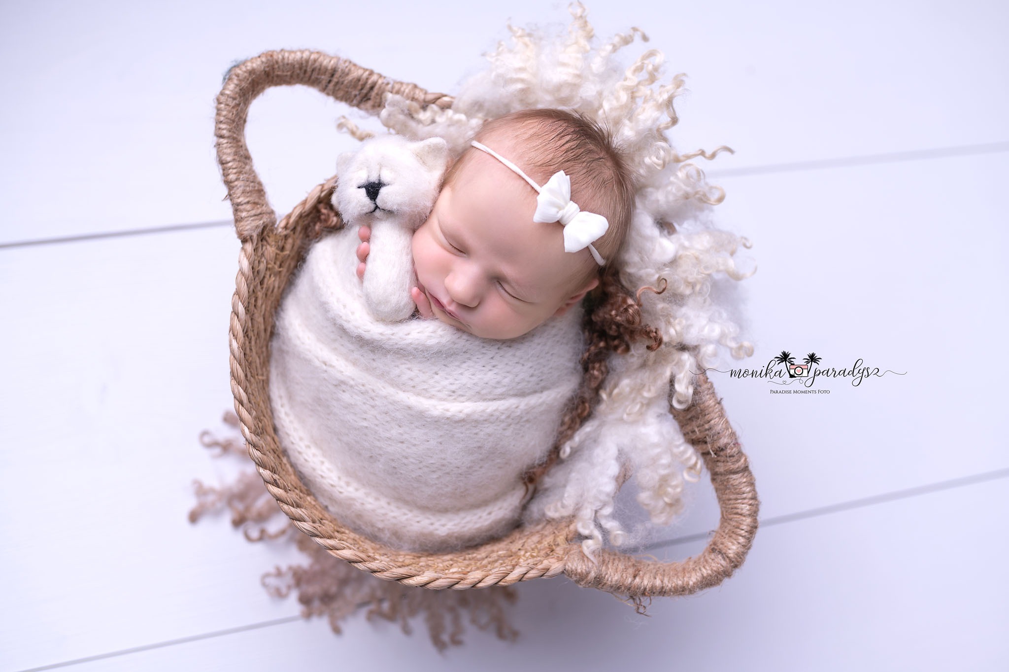 Noworodek zawinięty w kremowy kocyk, śpiący w wiklinowym koszyku z pluszowym misiem, białe tło.