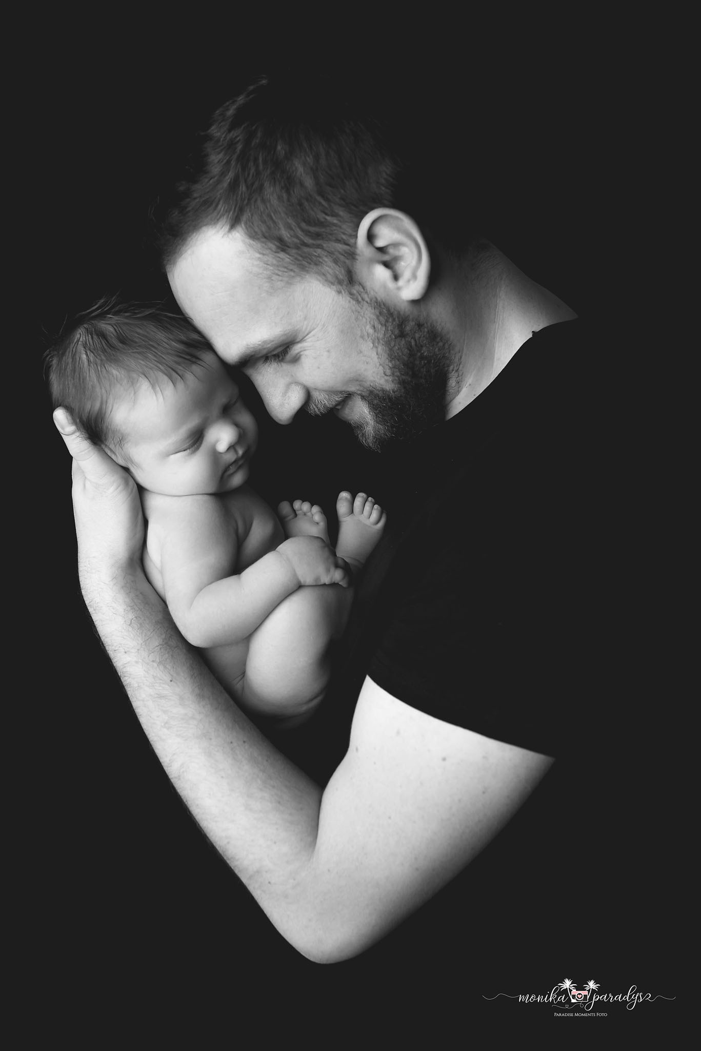 Czarno-białe zdjęcie ojca trzymającego śpiącego noworodka, z czułym spojrzeniem skierowanym w dół, na ciemnym tle.