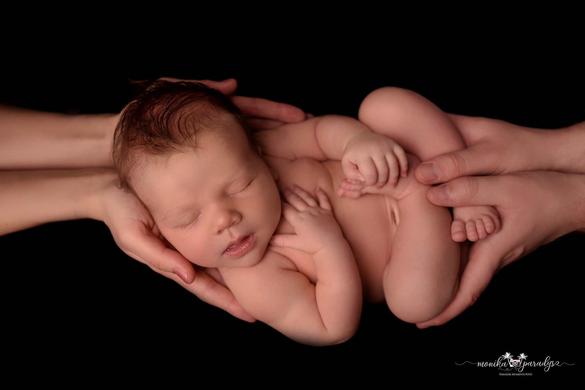 Noworodek śpiący w dłoniach rodziców na czarnym tle, delikatne oświetlenie podkreśla detale skóry i włosów dziecka.