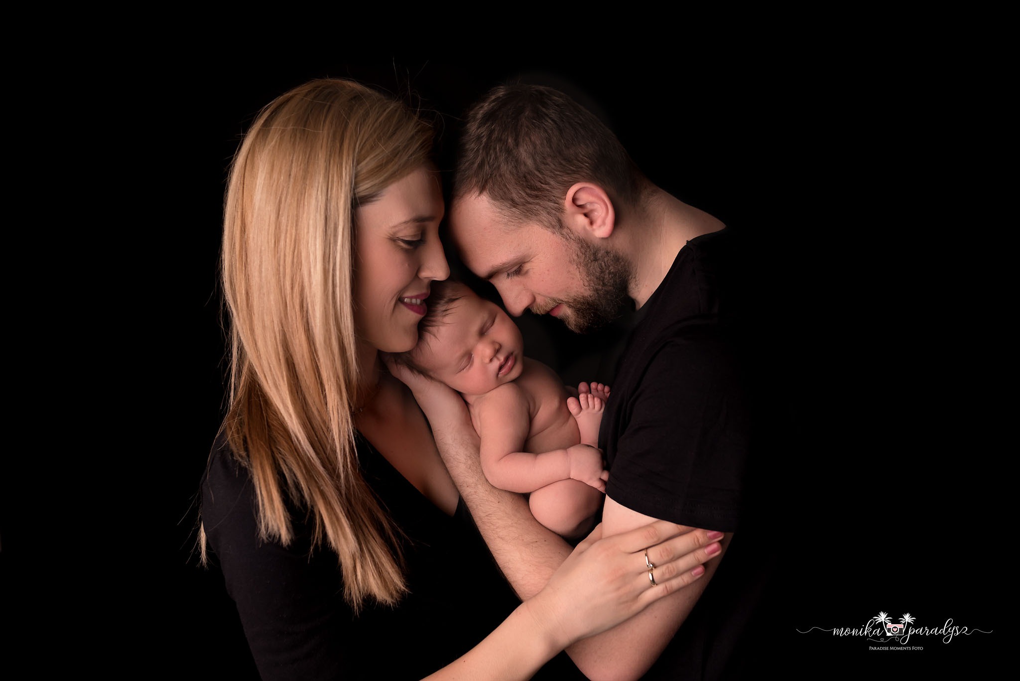 Rodzice trzymają śpiącego noworodka na czarnym tle, delikatne oświetlenie podkreśla bliskość i czułość.