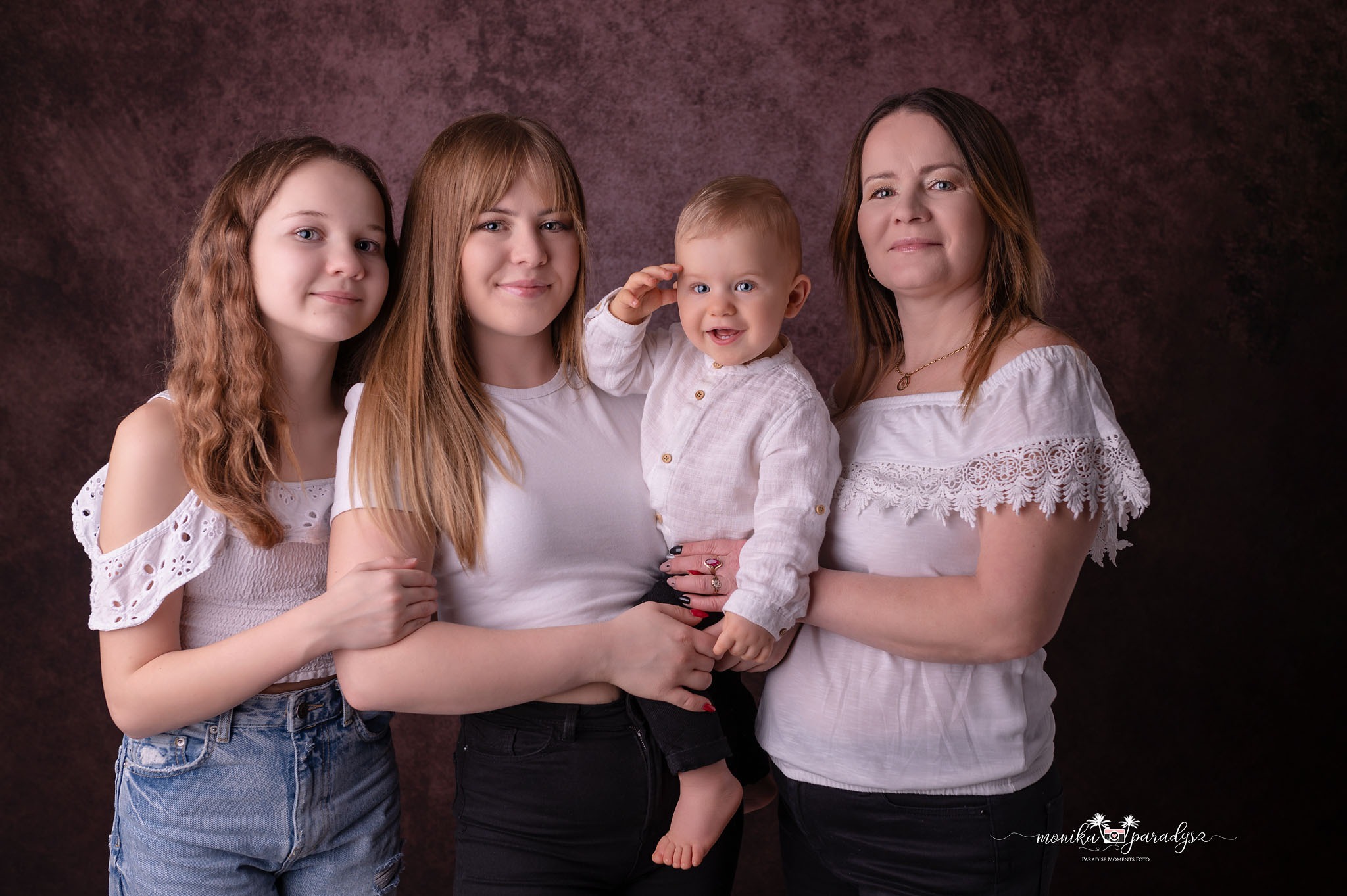 Portret rodzinny: mama, nastolatka, młoda kobieta i małe dziecko w białych ubraniach na ciemnym, teksturowanym tle. Dziecko trzyma rękę przy czole w geście pozdrowienia.