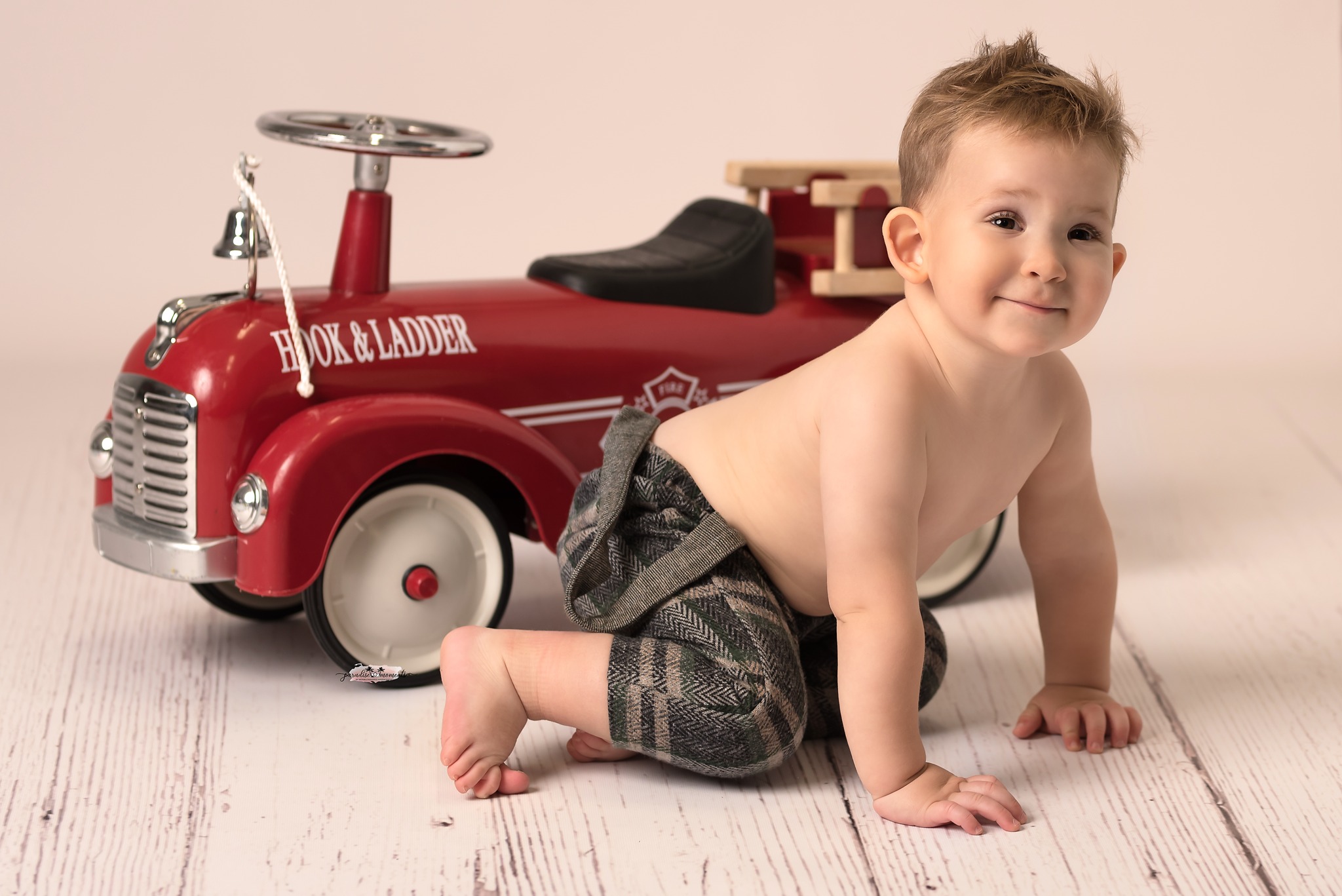 Uśmiechnięty niemowlak w tweedowych spodniach raczkuje obok czerwonej, metalowej zabawki wozu strażackiego Hook & Ladder na drewnianej podłodze.