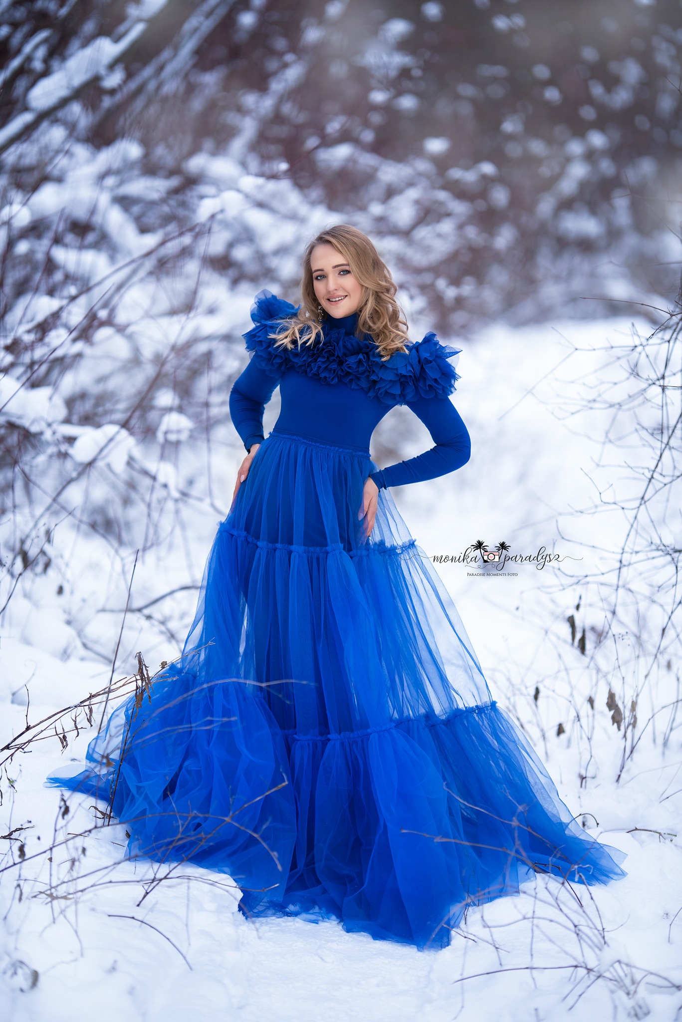 Kobieta w długiej, błękitnej sukni z falbanami na tle zimowego krajobrazu z zaspami śniegu i gałęziami drzew