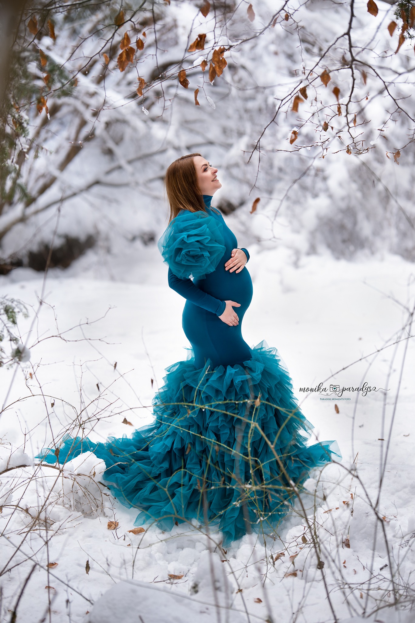 Kobieta w ciąży w długiej, turkusowej sukni z falbanami, pozuje w zasypanym śniegiem lesie, spoglądając w górę na gałęzie z brązowymi liśćmi.