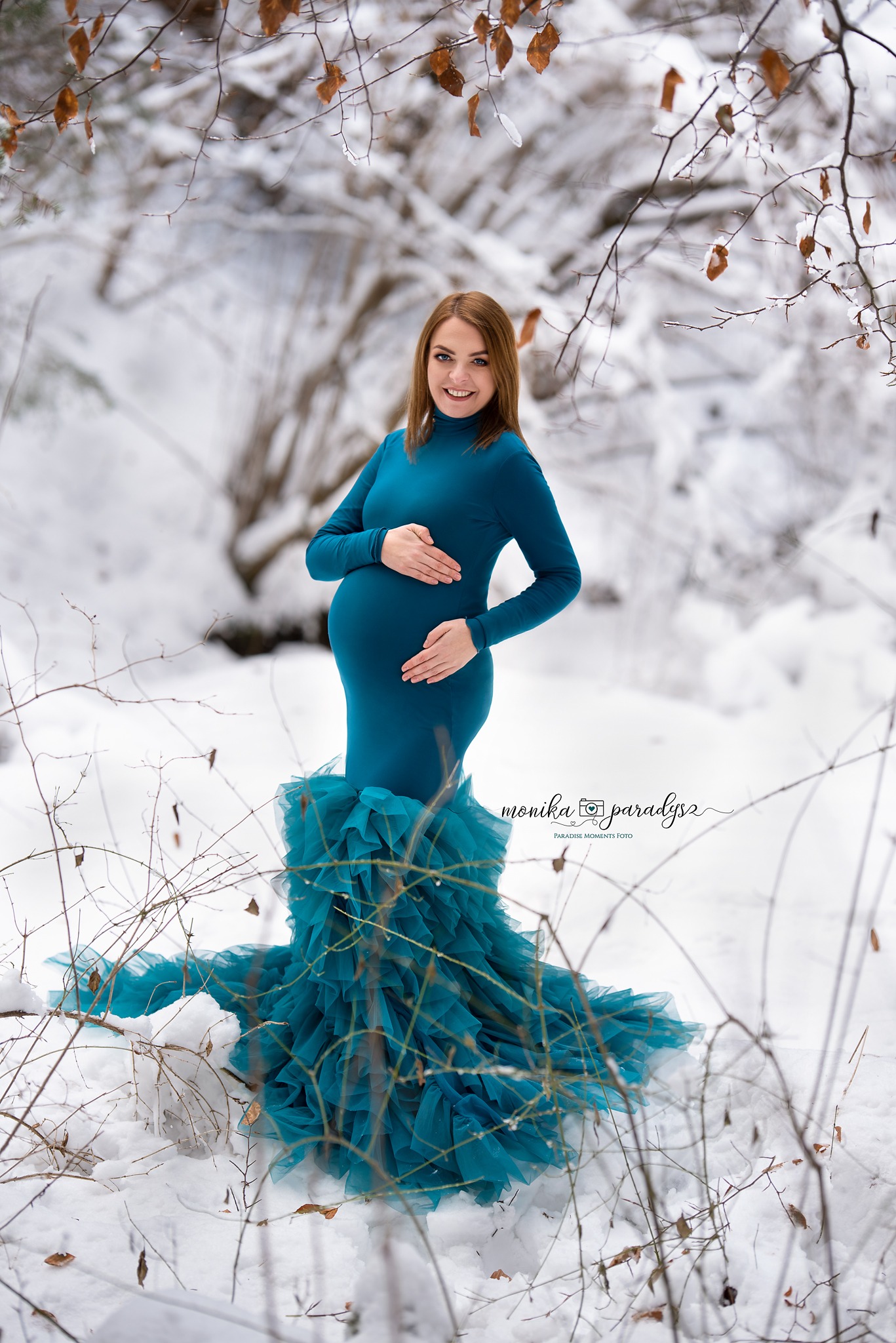 Uśmiechnięta, ciężarna kobieta w turkusowej sukni z falbanami pozuje na śniegu w zimowym lesie, trzymając ręce na brzuchu.
