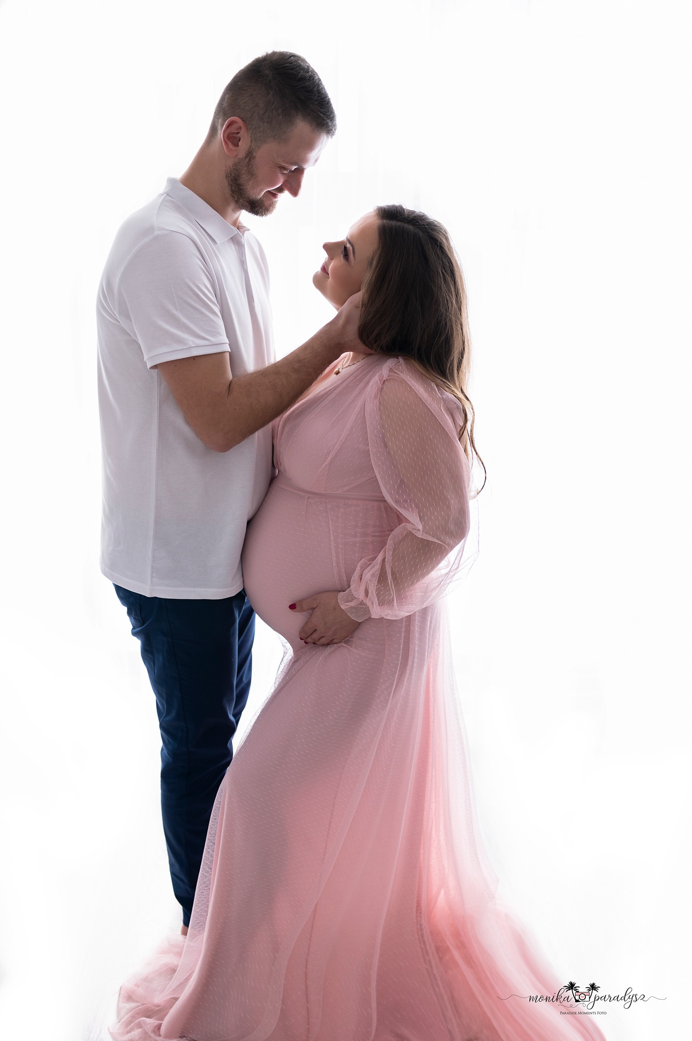 Przyszła mama w długiej, różowej sukni z tiulowymi rękawami, obejmowana przez partnera, patrzą na siebie na jasnym, studyjnym tle.