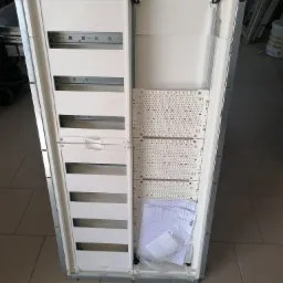 Otwarta, pusta rozdzielnica elektryczna z widocznymi szynami montażowymi i elementami mocującymi, leżąca na podłodze z dokumentacją i akcesoriami wewnątrz.