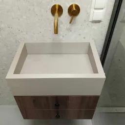 Nowoczesna, prostokątna umywalka z szarego betonu architektonicznego, zawieszona na szafce z drewnianymi szufladami o wzorze drewna, z złotą armaturą na tle ściany wykończonej mozaiką.