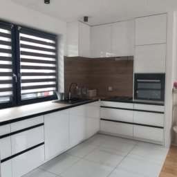 Parago Meble Tomasz Hańczaruk - Biała, nowoczesna kuchnia z czarnymi akcentami. Błyszczące fronty szafek, drewniany panel nad blatem, żaluzje w oknie. Minimalistyczny design.