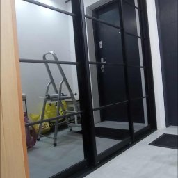 Parago Meble Tomasz Hańczaruk - Nowoczesne drzwi przesuwne z czarnymi ramami, widoczne w korytarzu z szarą podłogą. W tle drabina i torby.