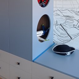 Parago Meble Tomasz Hańczaruk - Niebiesko-szara szafa z okrągłymi otworami, w których widać zabawki. Na blacie leży piracka czapka. Na ścianie rysunek statku i postaci.