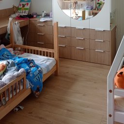 Parago Meble Tomasz Hańczaruk - Pokój dziecięcy z drewnianymi meblami: łóżko z barierkami, komoda z szufladami i szafa z lustrem w kształcie półkola. Na podłodze panele imitujące drewno.