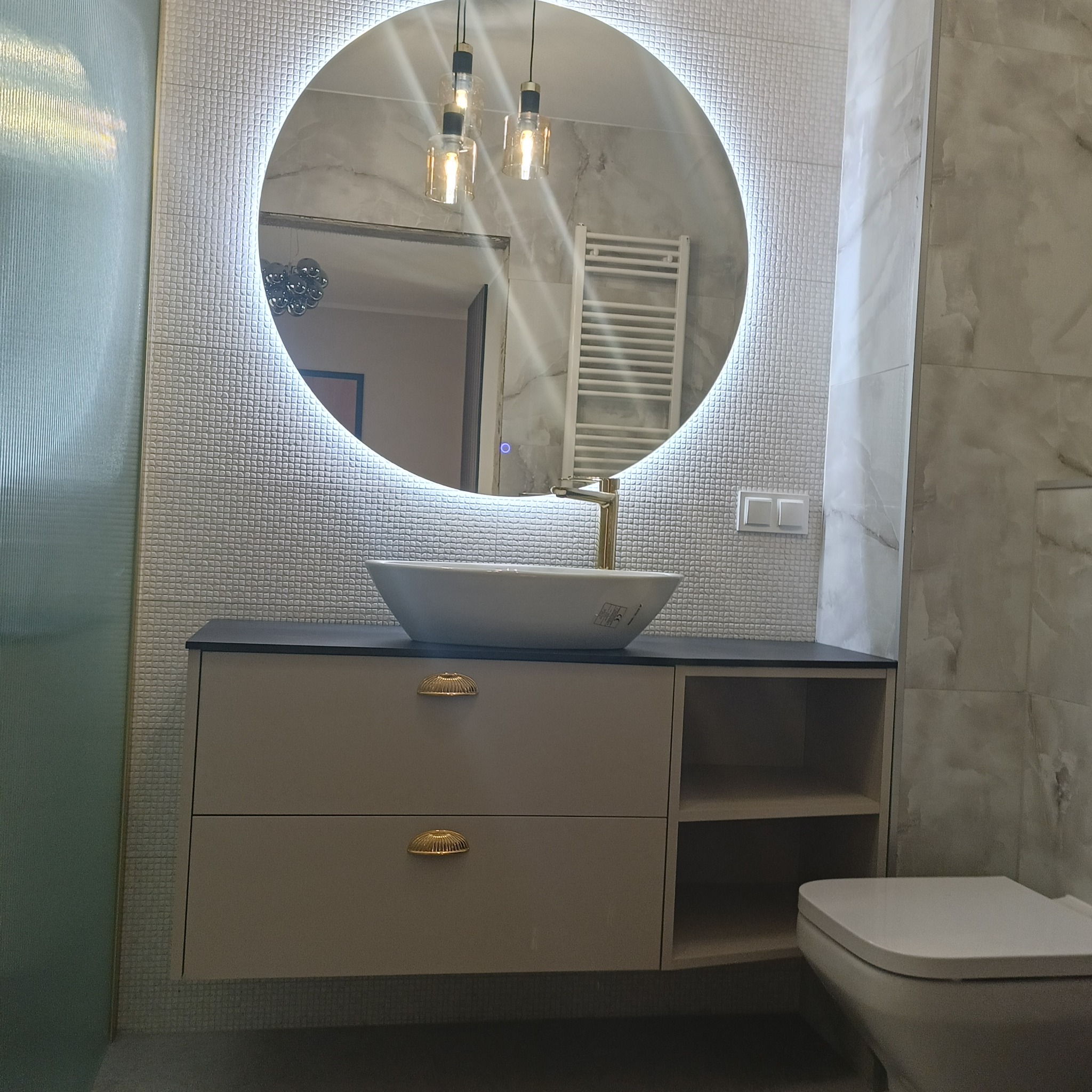 Nowoczesna łazienka z umywalką nablatową, szafką z szufladami z ozdobnymi uchwytami, okrągłym lustrem z podświetleniem LED i złotą baterią. W tle widoczny grzejnik drabinkowy.