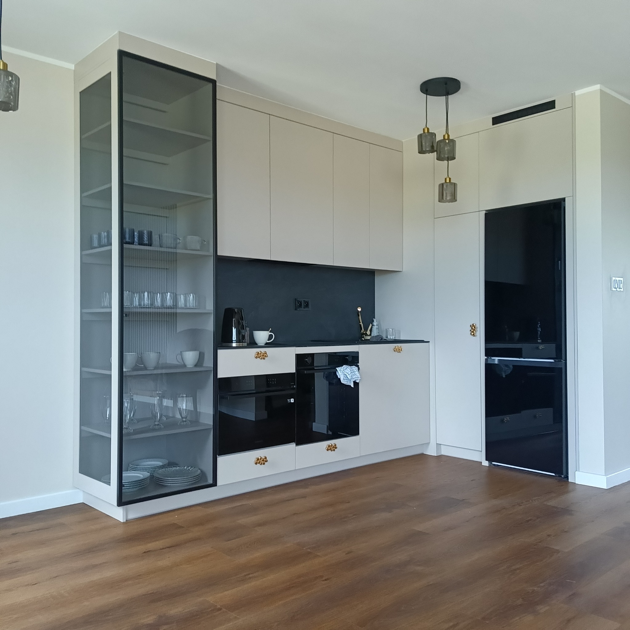 Nowoczesna kuchnia z beżowymi szafkami, czarnym panelem nad blatem i przeszkloną witryną, oświetlona wiszącymi lampami.