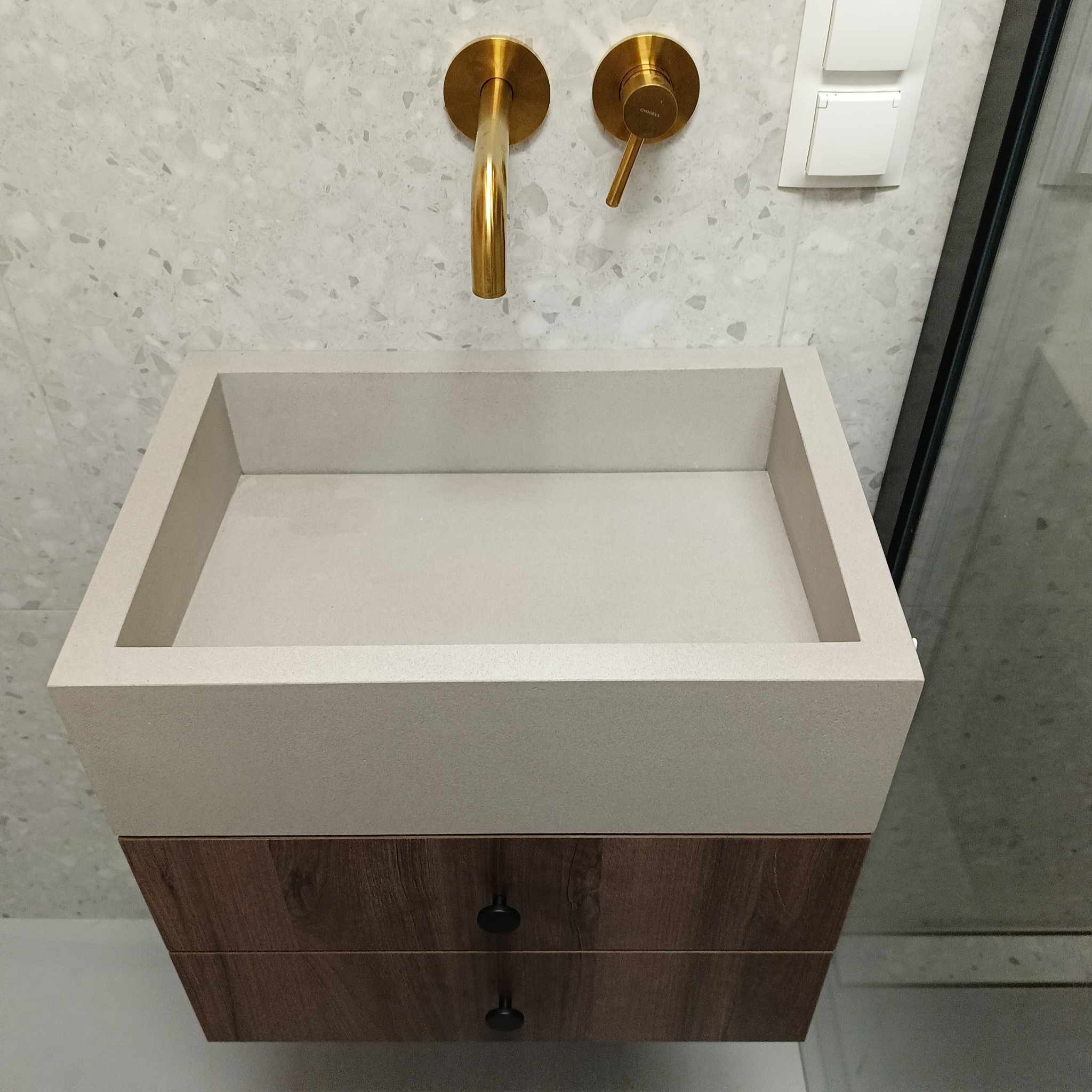 Nowoczesna, prostokątna umywalka z szarego betonu architektonicznego, zawieszona na szafce z drewnianymi szufladami o wzorze drewna, z złotą armaturą na tle ściany wykończonej mozaiką.