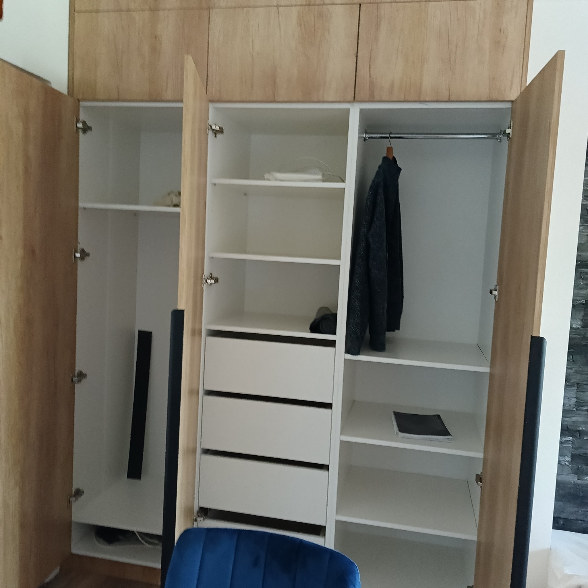Otwarta szafa wnękowa z frontami imitującymi drewno, widoczne półki, szuflady i drążek na ubrania, z elementami garderoby w środku.