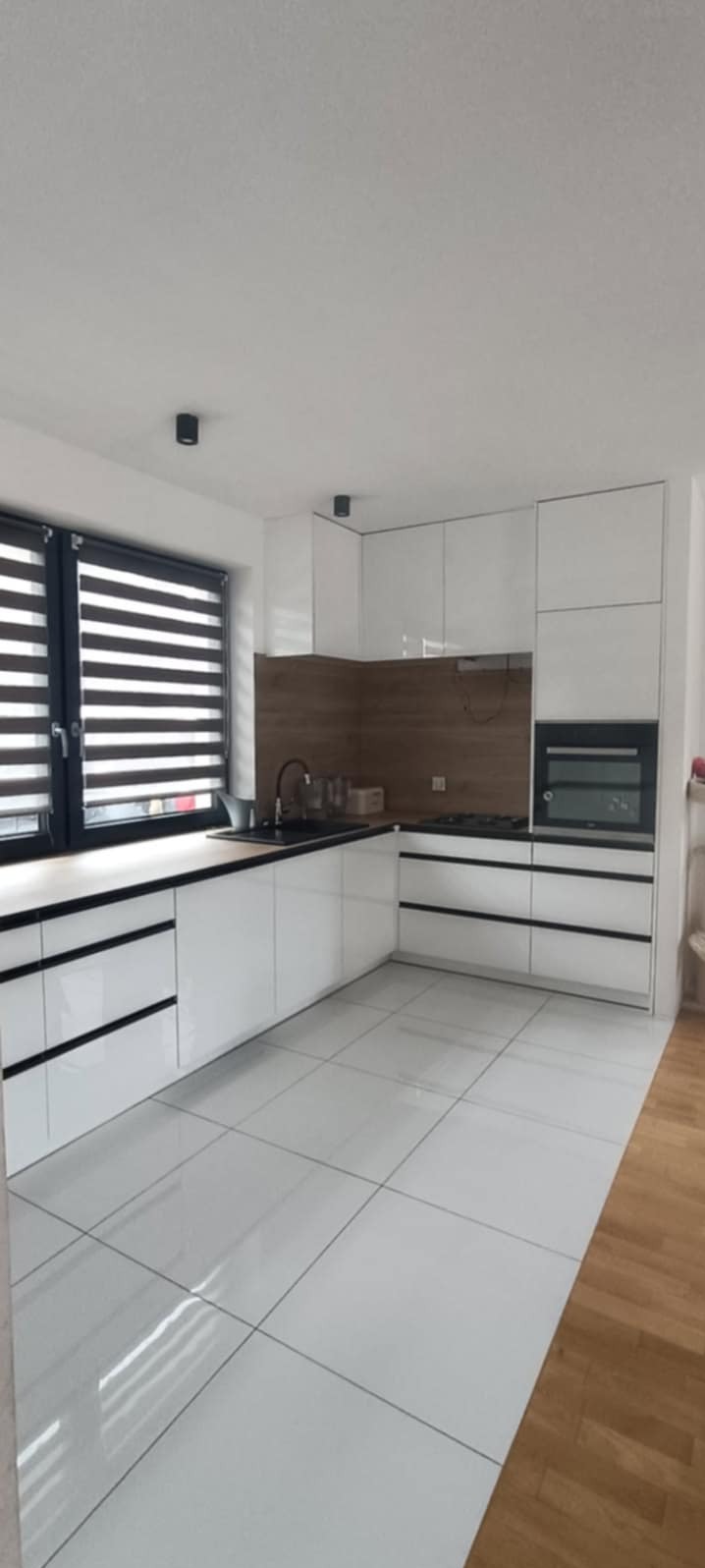 Biała, nowoczesna kuchnia z czarnymi akcentami. Błyszczące fronty szafek, drewniany panel nad blatem, żaluzje w oknie. Minimalistyczny design.