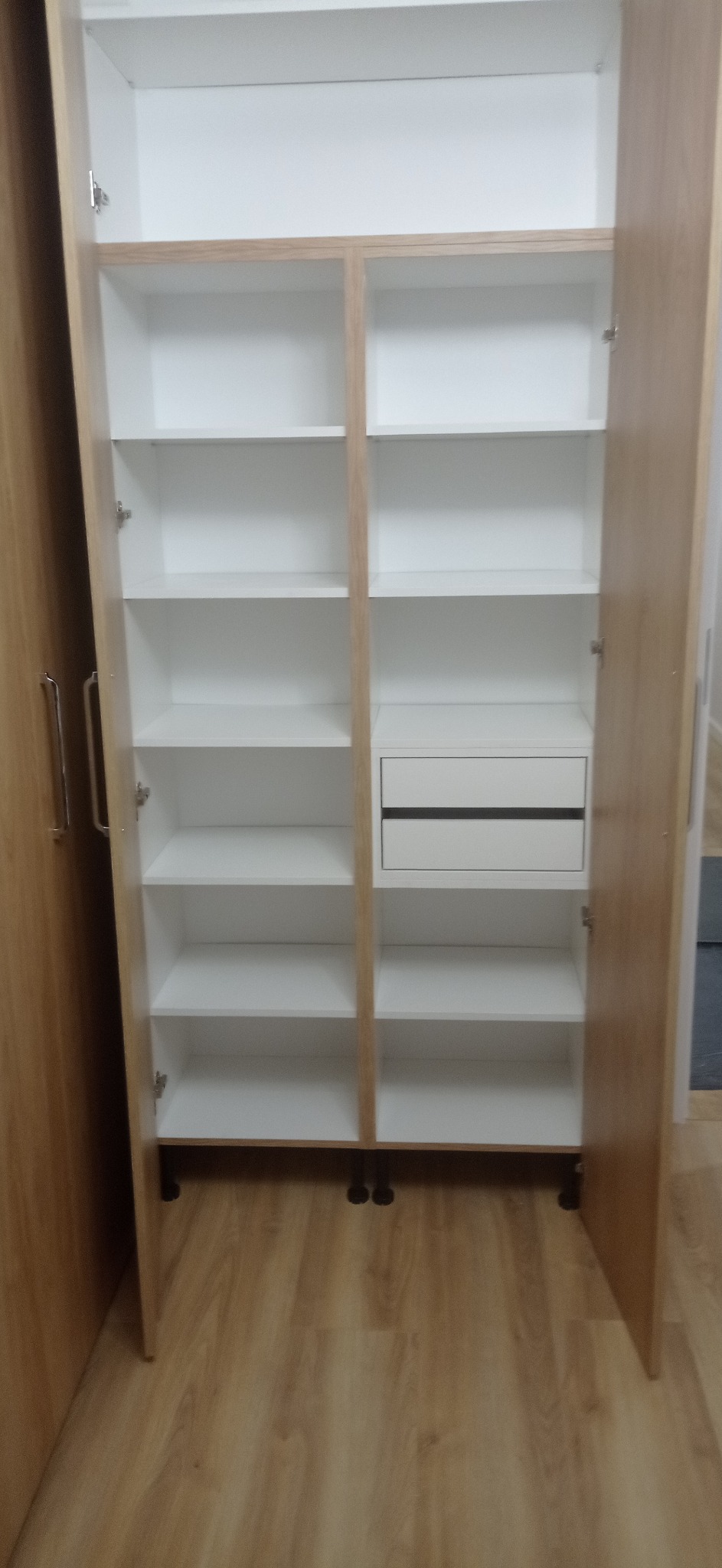 Wnętrze szafy wnękowej z półkami i szufladą, fronty w kolorze drewna, korpus biały, widok z otwartymi drzwiami, podłoga drewniana.