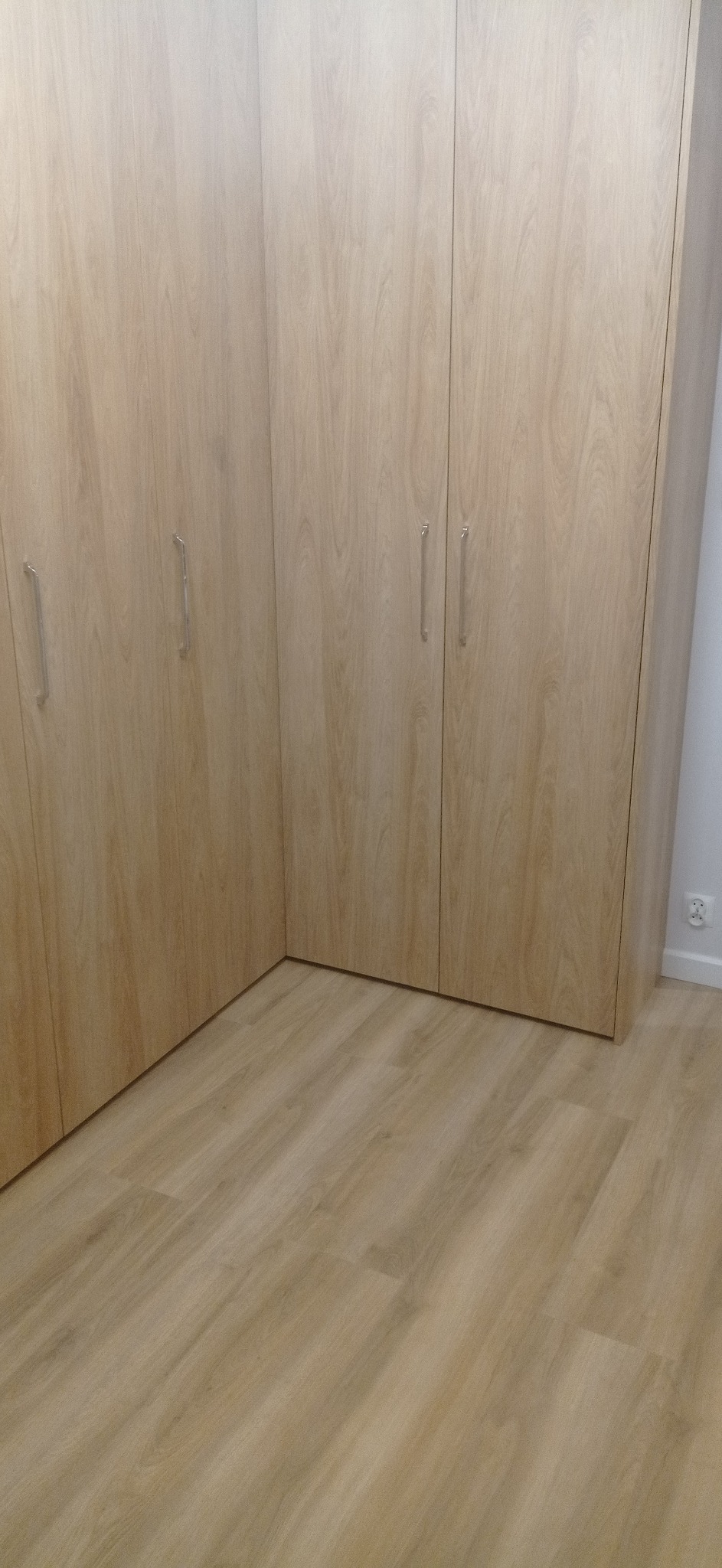 Jasna, narożna szafa wnękowa z drewnianymi frontami i metalowymi uchwytami, dopasowana do koloru podłogi. Widok na wnętrze szafy i podłogę.