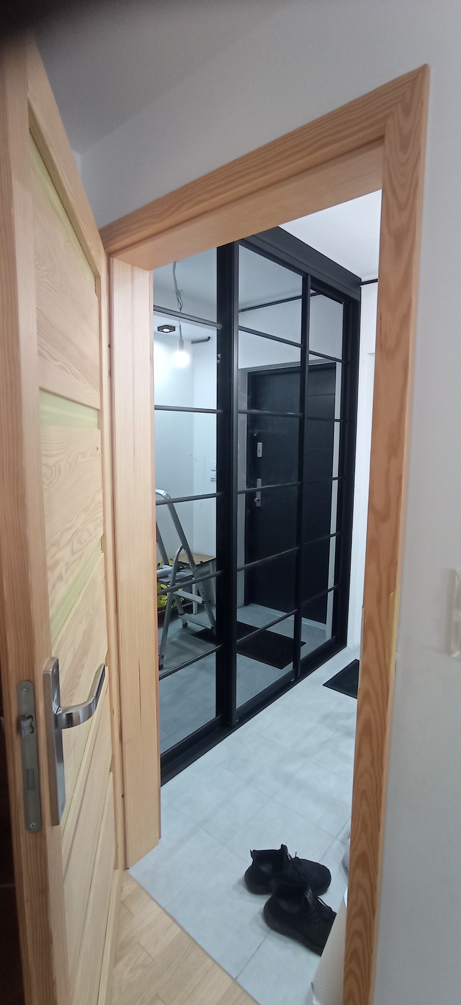 Otwarta drewniana rama drzwiowa z widokiem na przeszkloną, czarną szafę wnękową z drzwiami przesuwnymi, drabina w tle. Widoczne buty na podłodze.