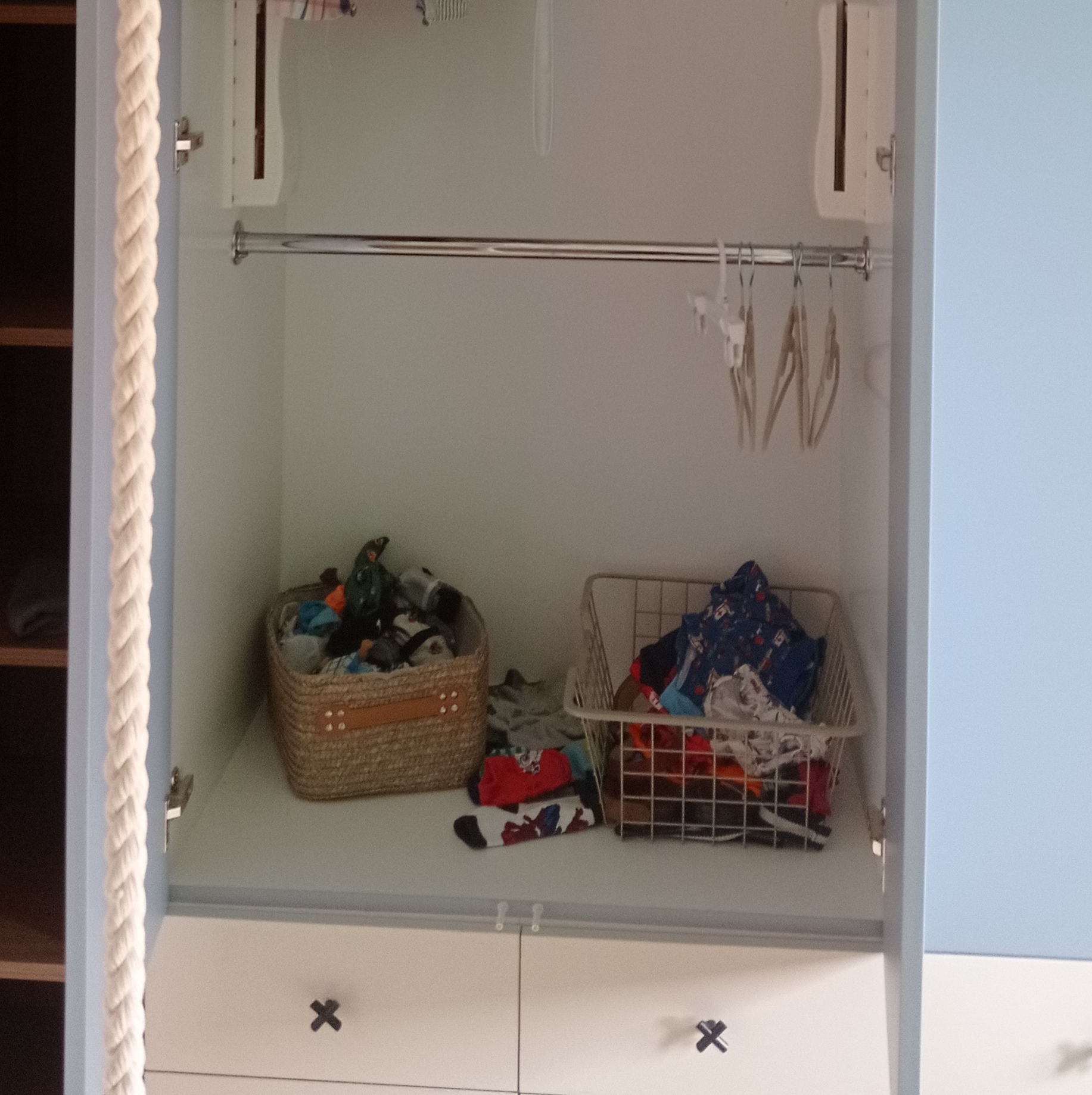 Otwarta szafa z jasnymi drzwiami, metalowym drążkiem na wieszaki i dwoma koszami z ubraniami na półce. Widoczne białe szuflady z czarnymi uchwytami w kształcie X.