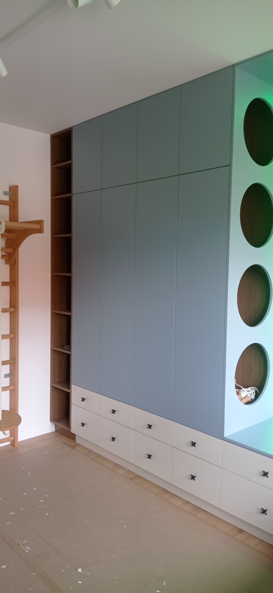 Niebiesko-biała szafa wnękowa z drewnianymi półkami i okrągłymi otworami z boku, czarne uchwyty, drabinka gimnastyczna z boku. Nowoczesny design.