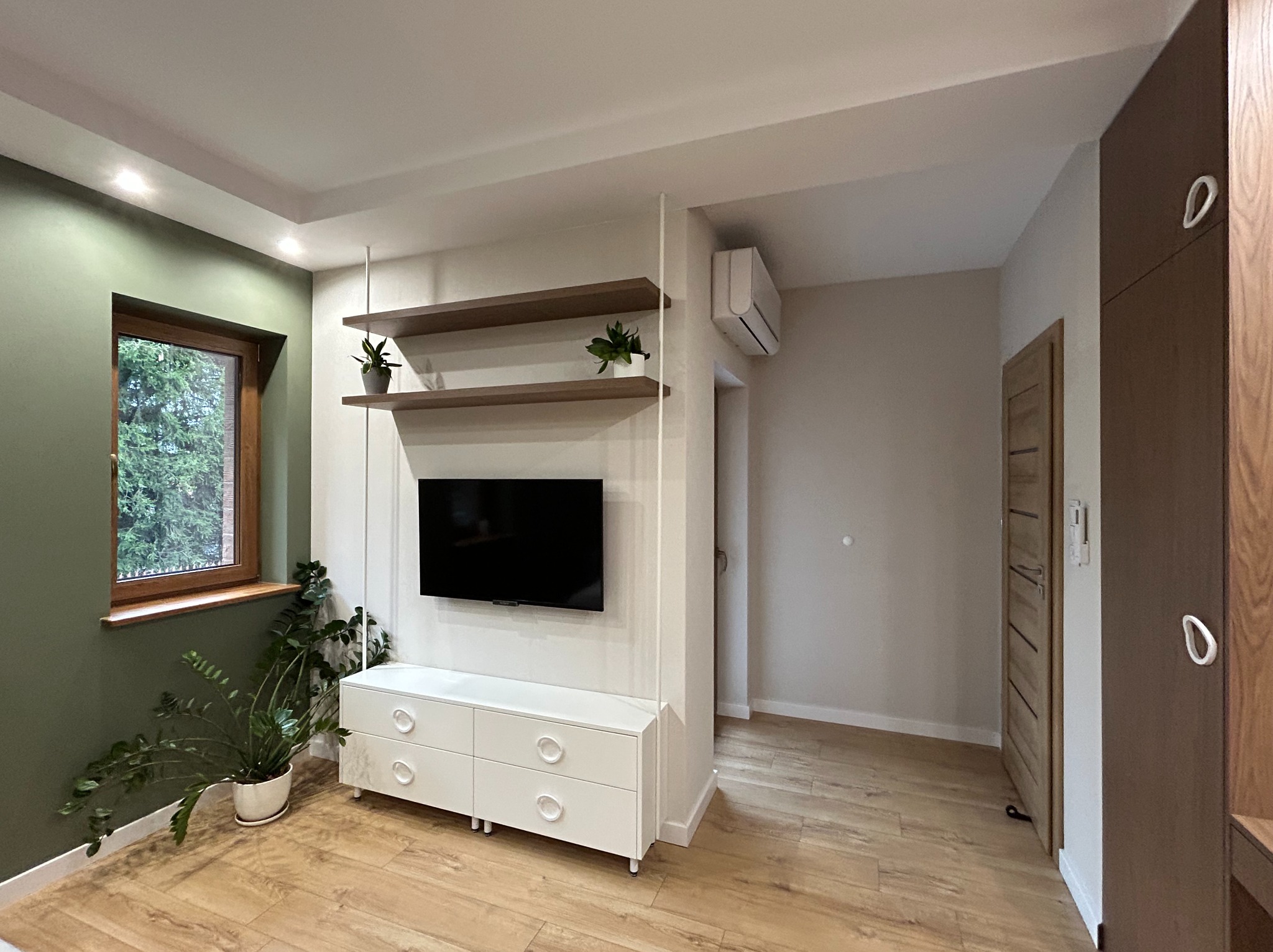 Nowoczesny salon z białymi meblami RTV, drewnianymi półkami i akcentami roślinnymi. Minimalistyczny design, ciepłe oświetlenie, drewniana podłoga i widok z okna.