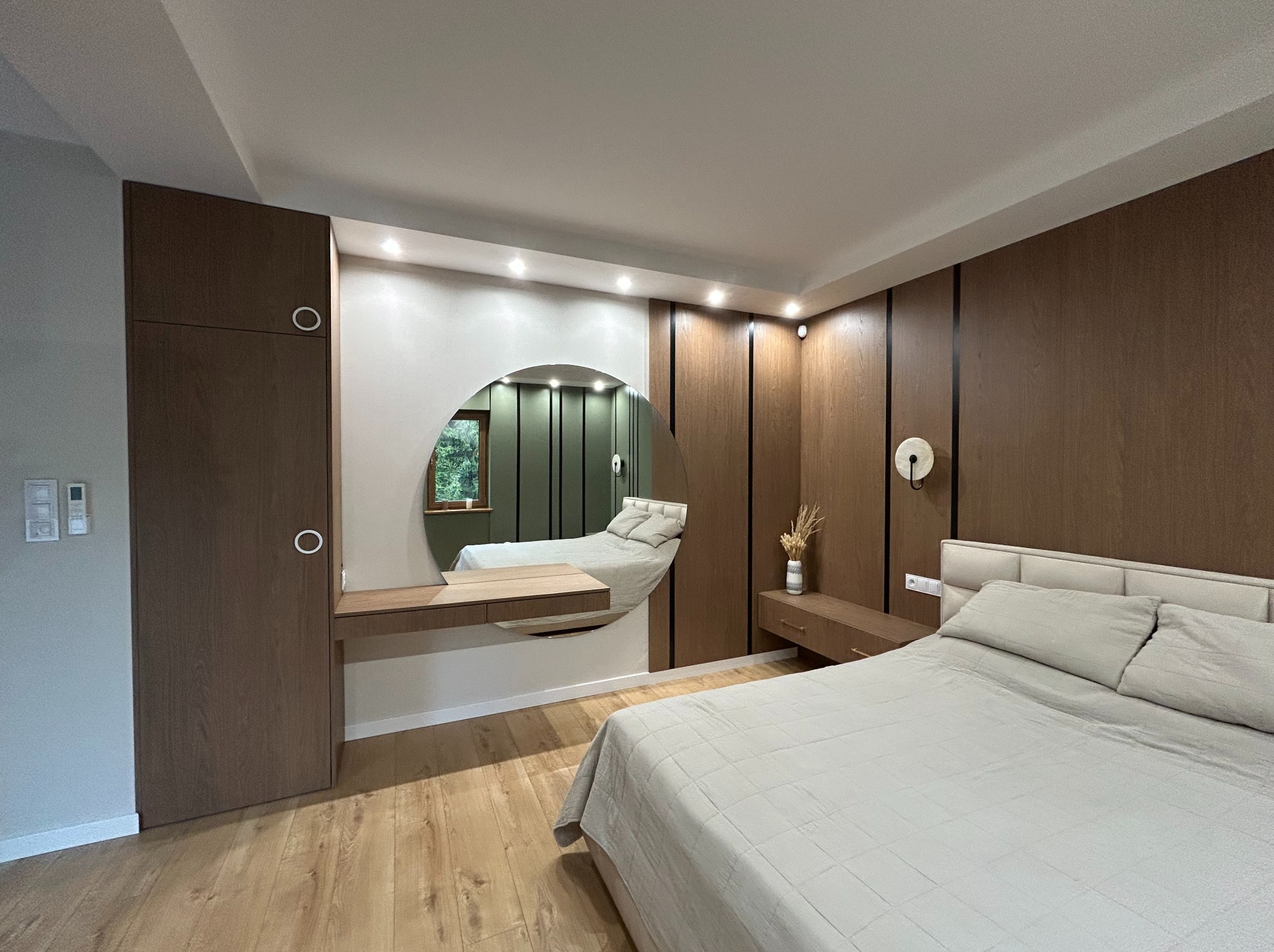 Nowoczesna sypialnia z zabudową drewnianą, okrągłym lustrem z toaletką i szafą wnękową. Uwagę przykuwa oryginalne oświetlenie i minimalistyczny design.