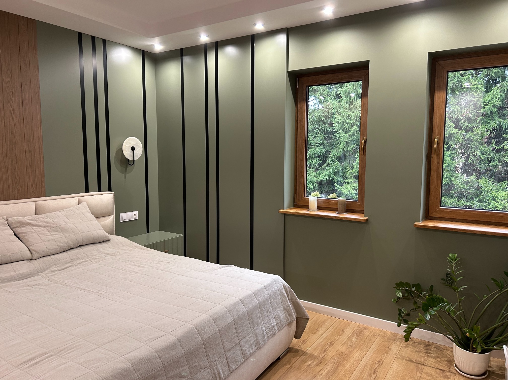 Nowoczesna sypialnia z łóżkiem, drewnianymi oknami i ścianą w odcieniach zieleni z czarnymi pasami. Widoczna lampa ścienna i roślina doniczkowa.