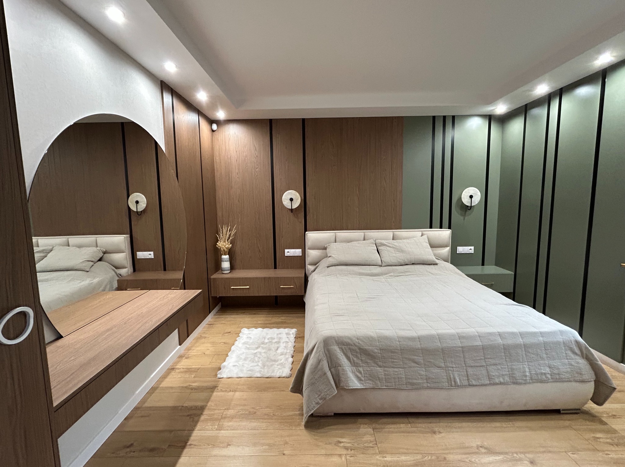 Nowoczesna sypialnia z łóżkiem, szafkami nocnymi i toaletką z lustrem w drewnianej okleinie. Minimalistyczne oświetlenie i dywanik dopełniają aranżację.