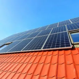 Panele fotowoltaiczne zamontowane na dachu z czerwoną dachówką, widoczne okna dachowe, błękitne niebo w tle, ujęcie z dołu.