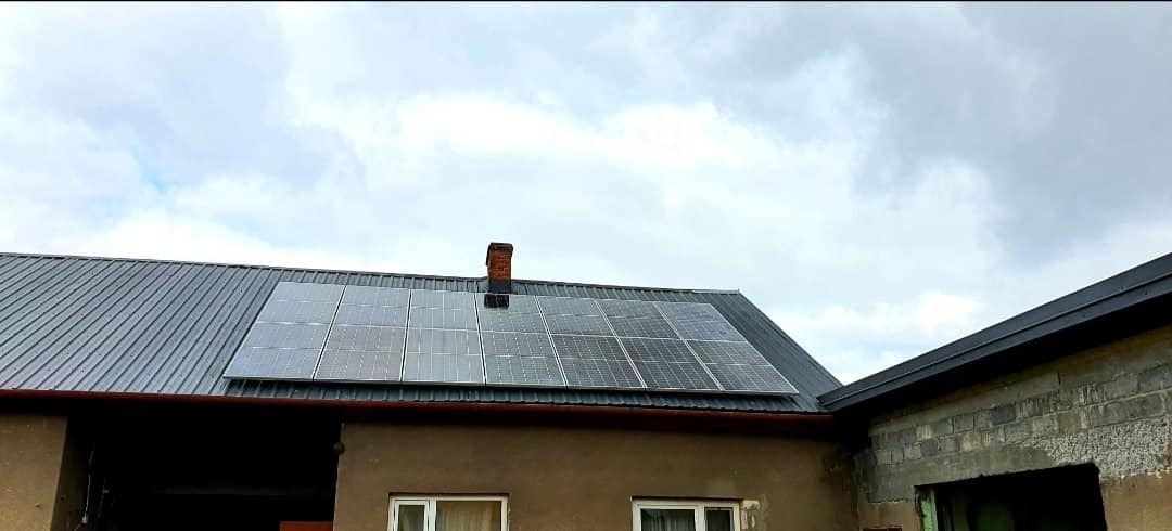 Panele fotowoltaiczne zamontowane na metalowym dachu domu z ceglanym kominem, widok z dołu w pochmurny dzień.