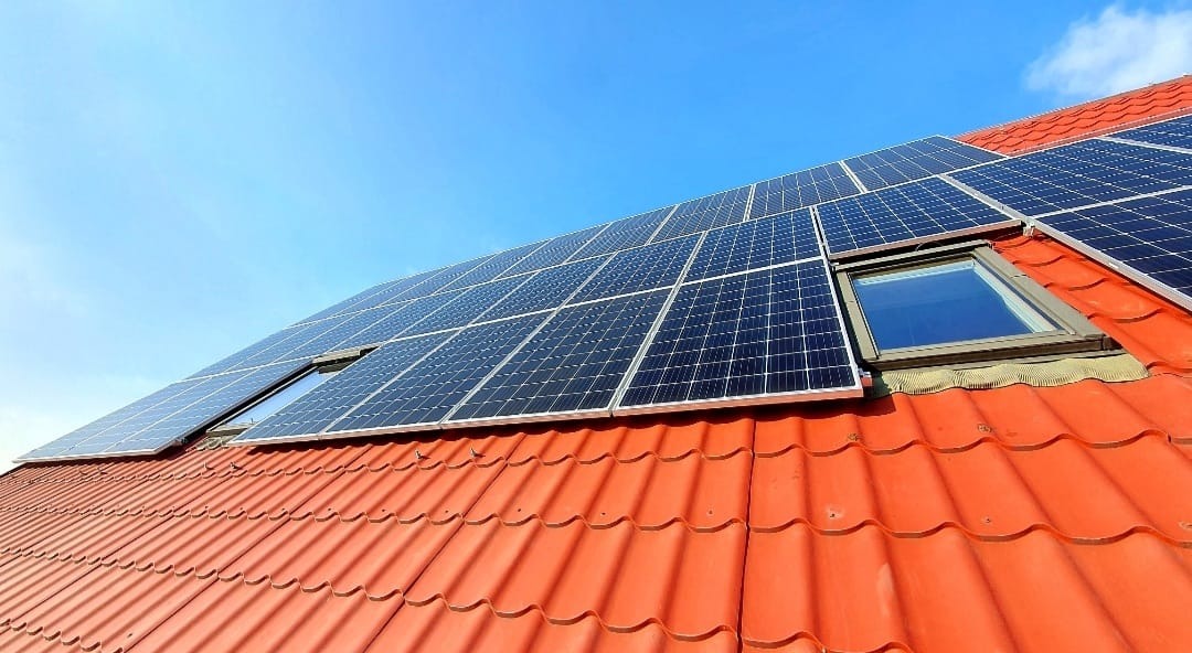 Panele fotowoltaiczne zamontowane na dachu z czerwoną dachówką, widoczne okna dachowe, błękitne niebo w tle, ujęcie z dołu.