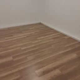 Ukończona podłoga z paneli laminowanych w odcieniach brązu, ułożona w rogu pokoju z białymi ścianami i listwami przypodłogowymi.